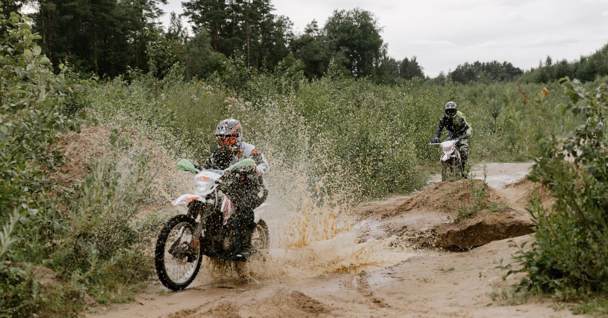 Two motorcyclists navigate a challenging muddy trail in a forest setting, showcasing extreme sports skills.